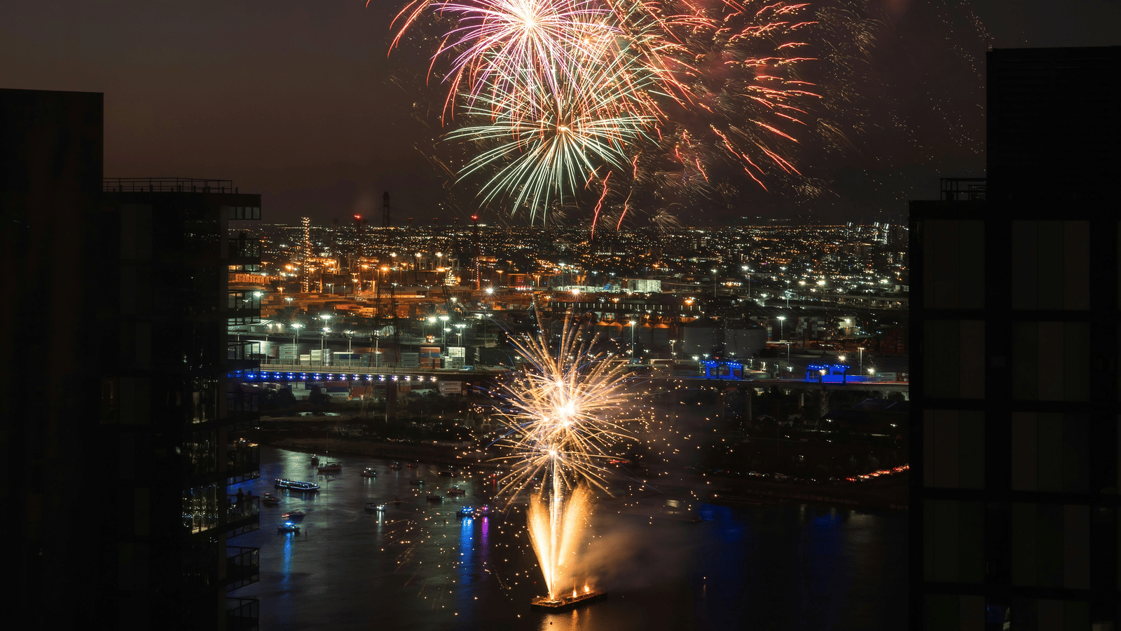 A night-time panoramic view of a city over a body of water with vibrant fireworks exploding in the sky.