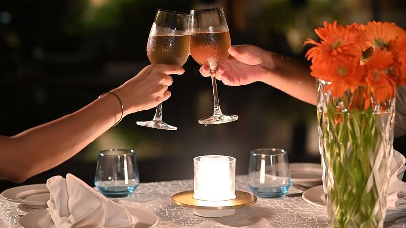 Close-up of a romantic candlelight dinner setup with two people clinking wine glasses, a bouquet of orange flowers, and gourmet appetisers on a white tablecloth - Ananta Spa and Resort, Pushkar.