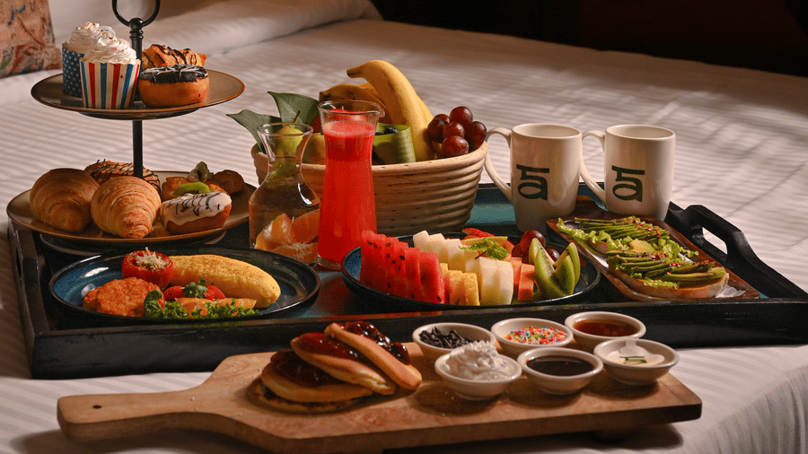 Luxurious breakfast spread served on a bed in a warmly lit hotel room, featuring a variety of dishes including fresh fruit, pastries, juice, and coffee - Ananta Spa and Resort, Pushkar.