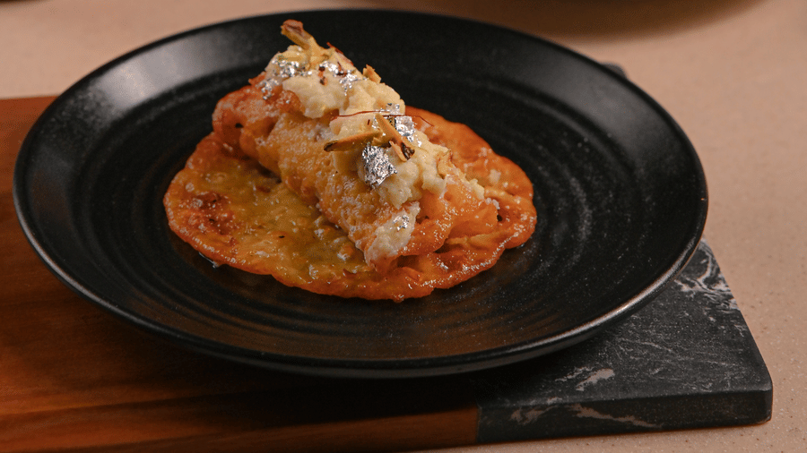 Close-up of a plated dish featuring a savoury item topped with sauce and herbs, with another similar dish in the background being eaten by a person, set on a table with a potted plant - Ananta Spa and Resort, Pushkar.