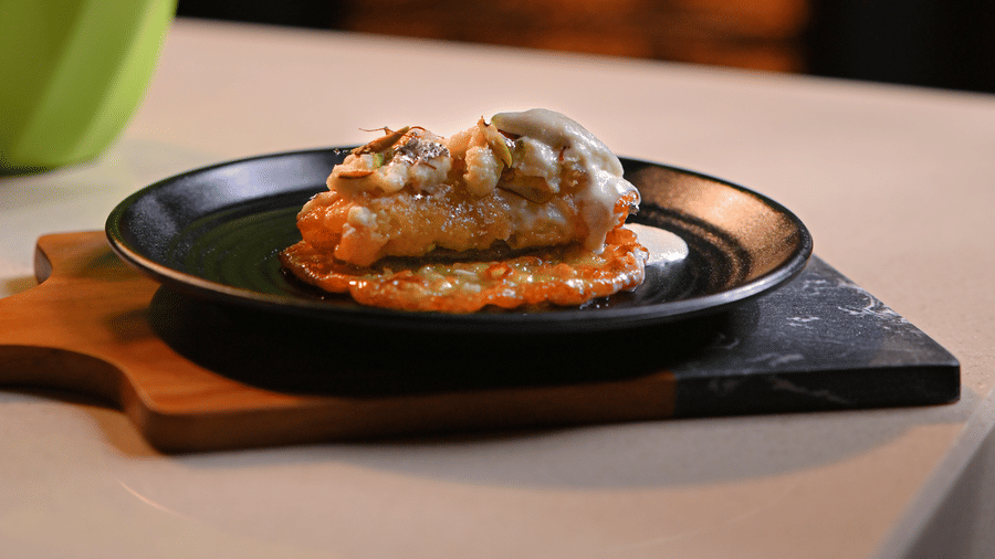 Close-up of a gourmet dish on a black plate with sauce being poured over it from a silver gravy boat, placed on a wooden serving board next to a potted green plant - Ananta Spa and Resort, Pushkar.