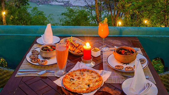 A candlelit outdoor dining table laden with food overlooking the river at Neemrana's Glasshouse on The Ganges - 21st Century, Rishikesh.