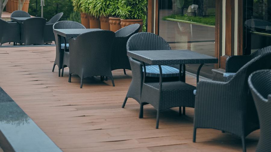 Outdoor seating area with wicker chairs and round tables on a wooden deck, adjacent to a building with a sloped tiled roof, surrounded by lush greenery and a blue umbrella in the background - Ananta Spa and Resort, Pushkar.
