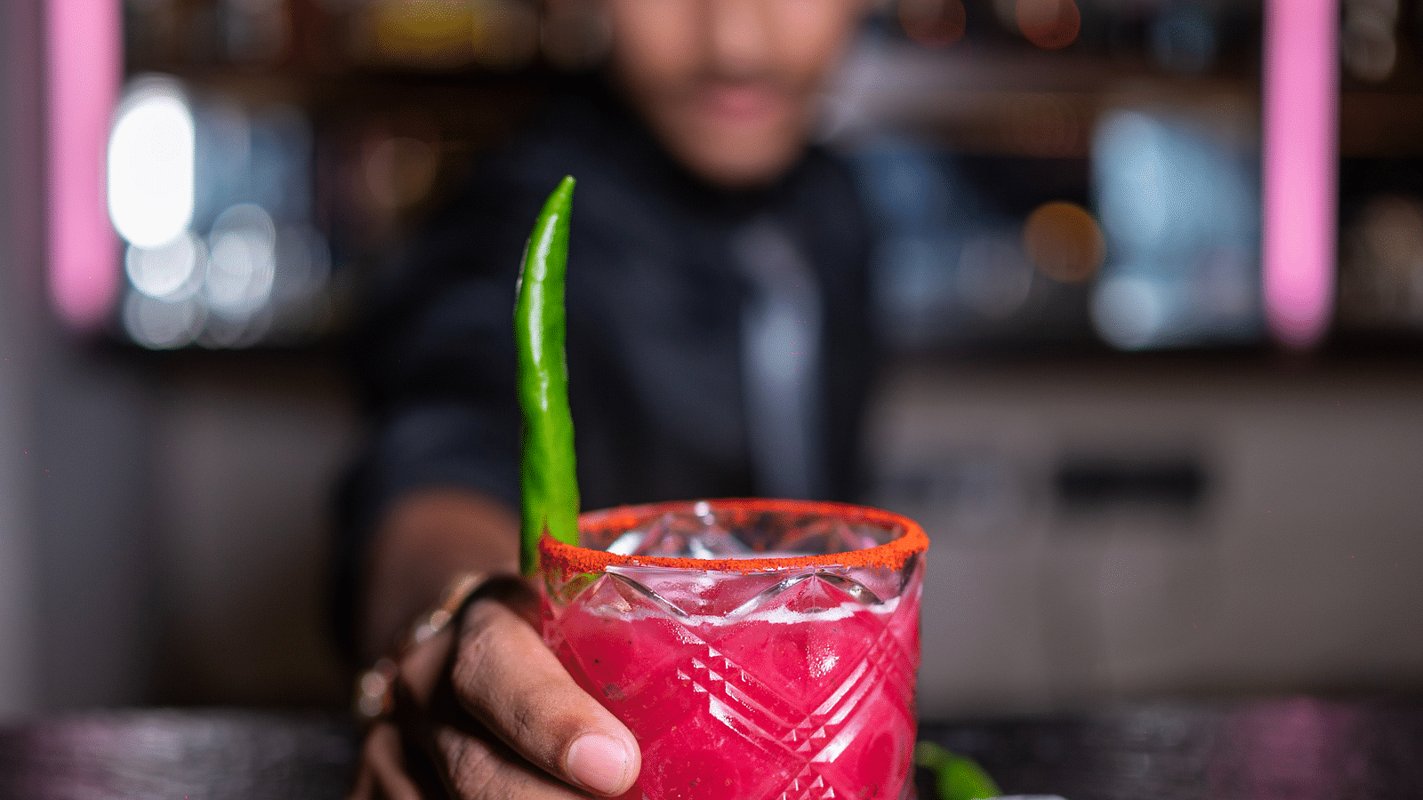 A bartender holding a drink garnished with a green chili, with the bartender blurred in the background at Hotel Moti Palace