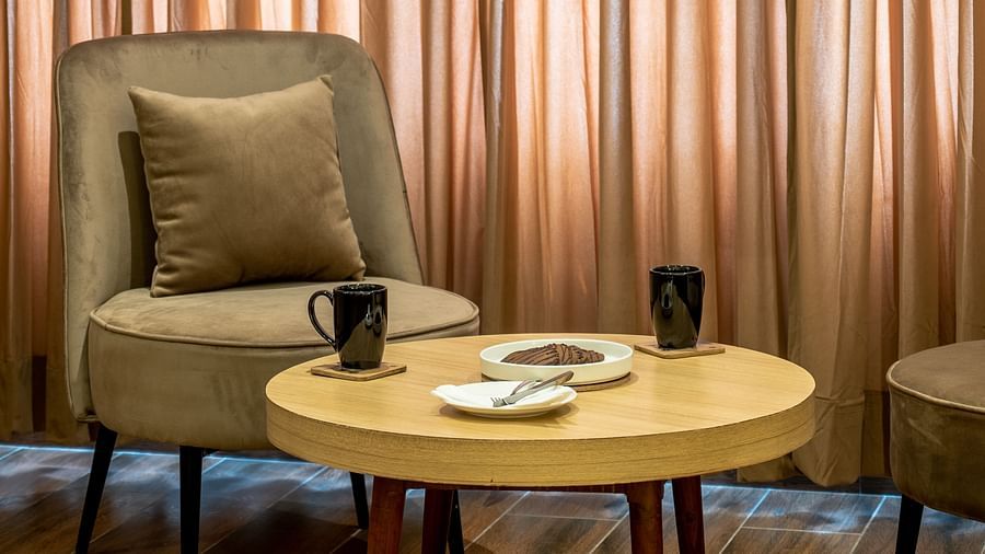 A living area of a suite at Vishranti Hotel Dehradun featuring a cushioned chair, a round table and a large window with curtains closed.