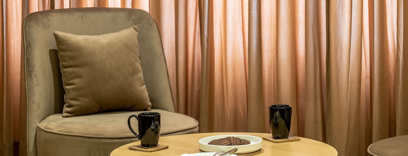 A living area of a suite at Vishranti Hotel Dehradun featuring a cushioned chair, a round table and a large window with curtains closed.