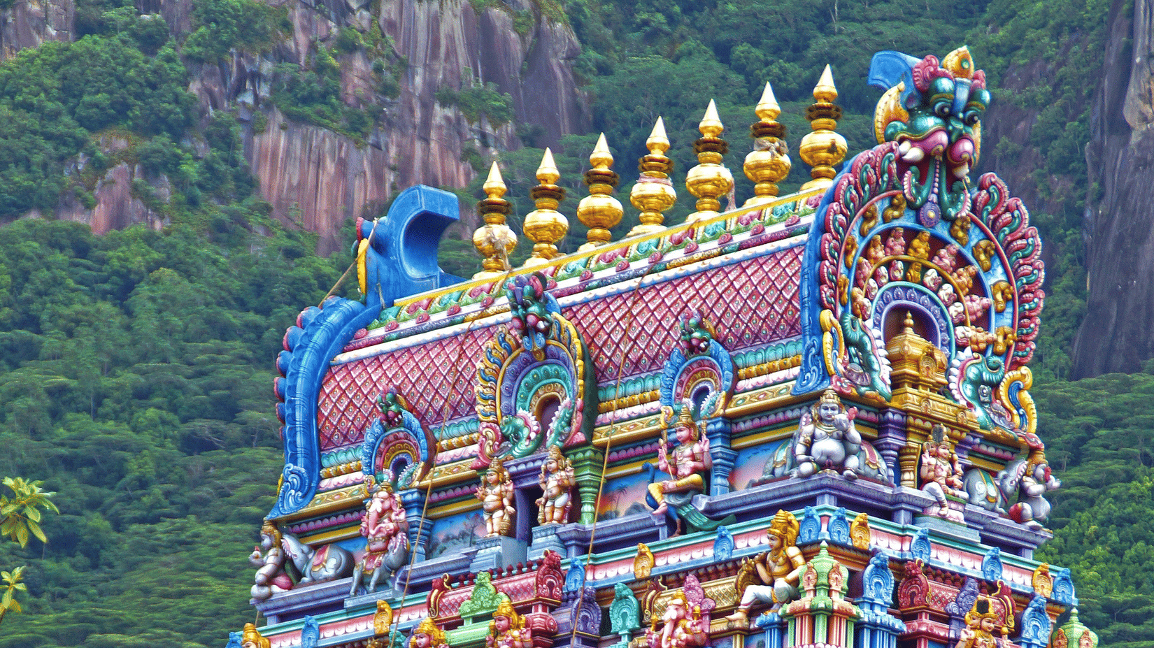 Tall, multi-tiered colourful gopuram tower of a traditional South Indian Hindu temple set against a green hill backdrop.