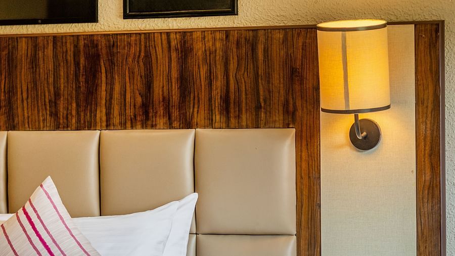 A close-up of a hotel bed with white linens and striped pillows, a wooden headboard, framed artwork on the wall, and a bedside lamp - Mango Hill Resort, Coimbatore
