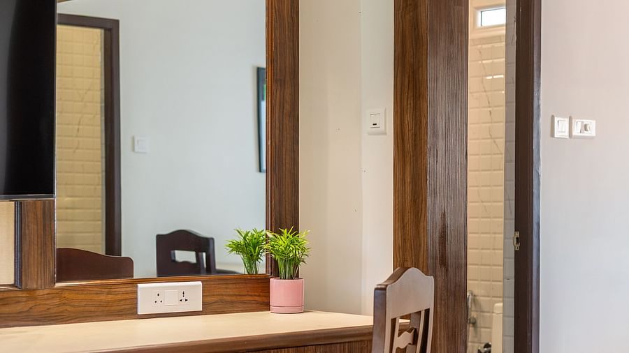 A hotel room with a wooden dresser, a large mirror, a chair, and a doorway leading to a bathroom - Mango Hill Resort, Coimbatore