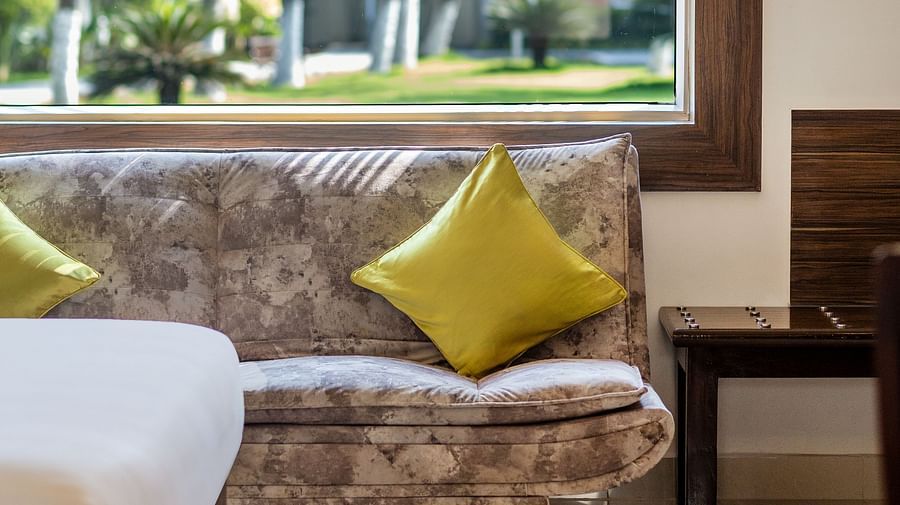 A patterned sofa with yellow pillows in a hotel room, with a window overlooking a garden with palm trees - Mango Hill Resort, Coimbatore