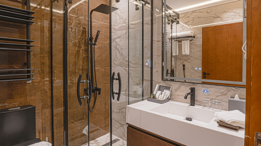 Bathroom of Standard Twin Room at Grand Iva Hotel, Abha, featuring a separate showering area, a wall-mounted mirror, sink, western commode, and marble flooring.