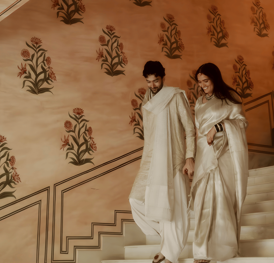 Charvi & Dhruv walking down staircase at Umaid Palace with pastel walls and traditional decor during wedding celebration.