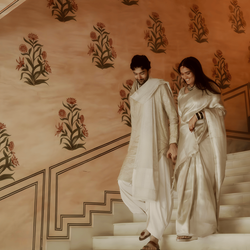 Charvi & Dhruv walking down staircase at Umaid Palace with pastel walls and traditional decor during wedding celebration.