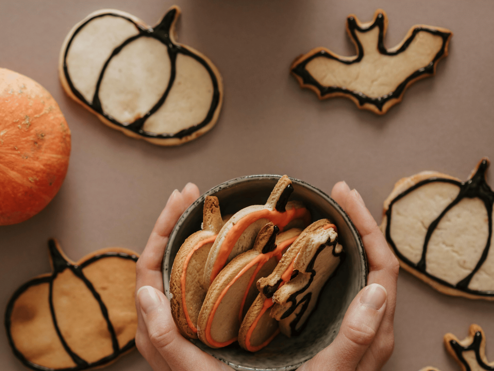 Hands holding small pumpkins surrounded by Halloween-themed cookies shaped like bats and ghosts.