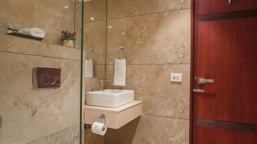 A hotel bathroom with marble-effect tiling, a wall-mounted toilet, a square basin, a dark wood door, and a large mirror at BluSalzz Select City Centre, Amritsar.