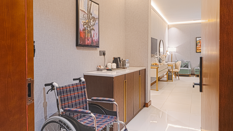 A wheelchair placed in the entrance of the ADA-friendly Deluxe King Rooms, as the door opens to a bright lit interior, and a cabinet | Grand Iva Hotel, Abha