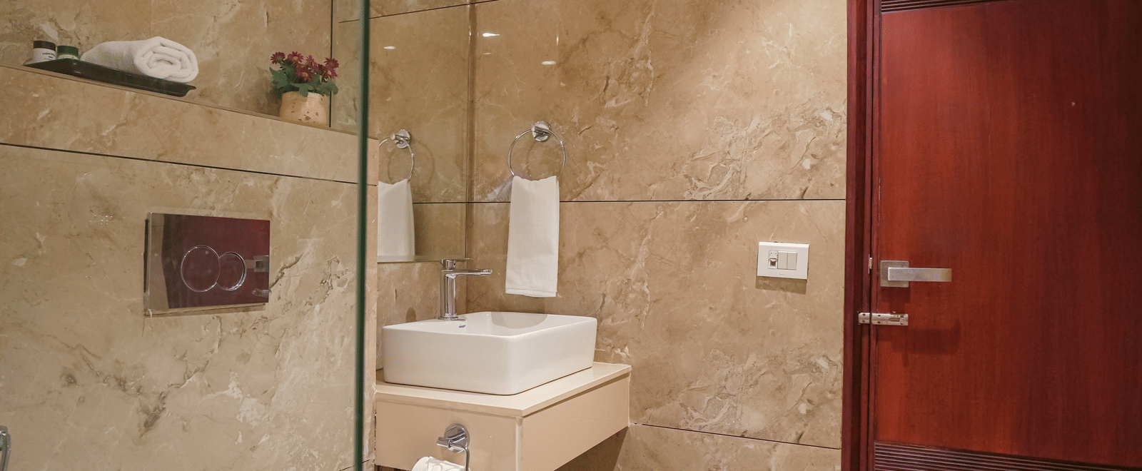 A hotel bathroom with marble-effect tiling, a wall-mounted toilet, a square basin, a dark wood door, and a large mirror at BluSalzz Select City Centre, Amritsar.