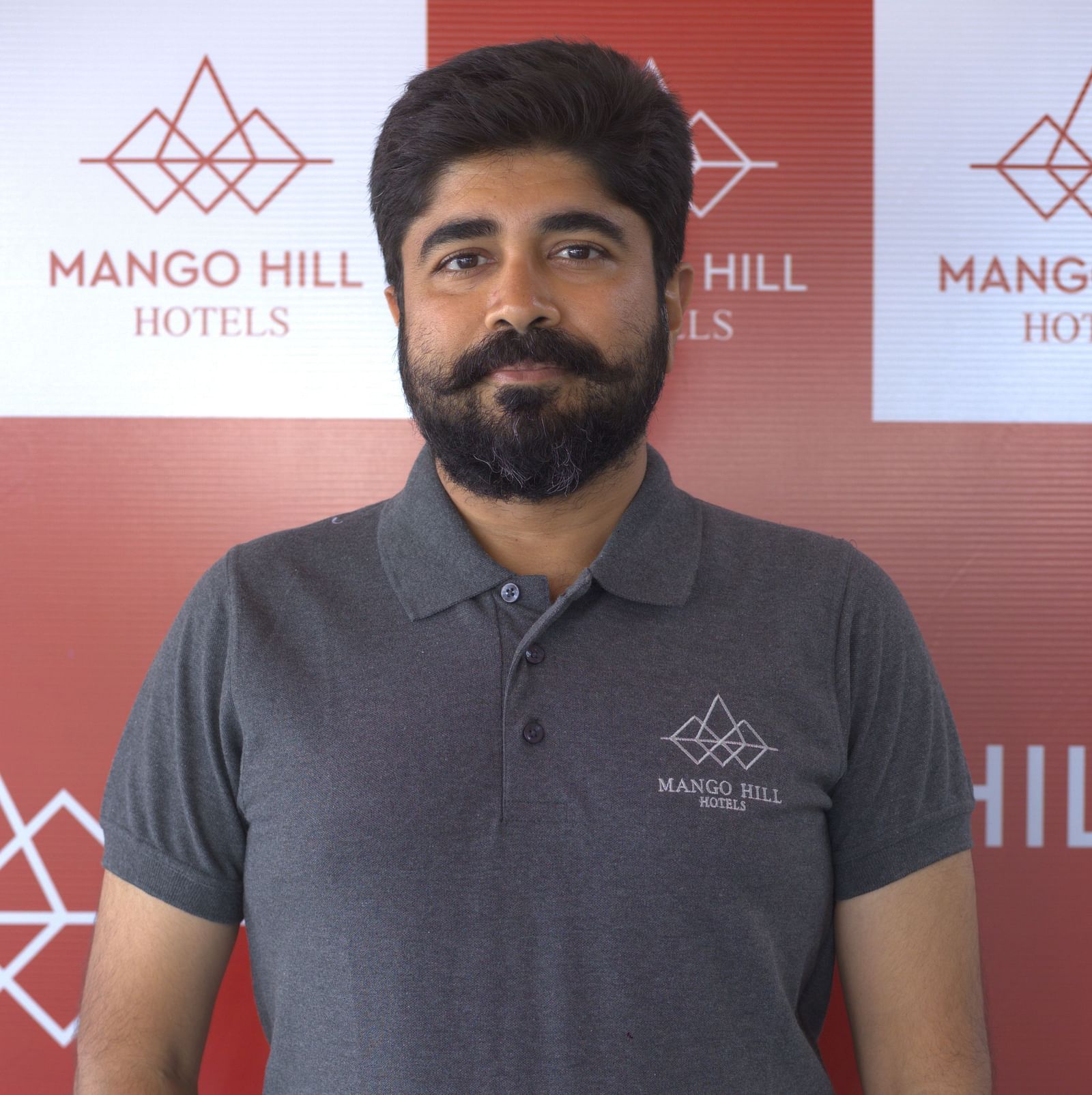 Om Saravanan in a grey Mango Hill Hotels uniform posing with hands in pockets in front of a branded backdrop. 