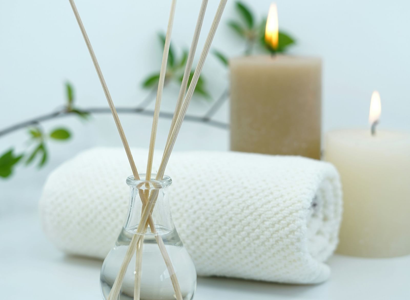 An arrangement of spa or wellness items, including a reed diffuser, a lit candle, and rolled white towels, set against a light background.