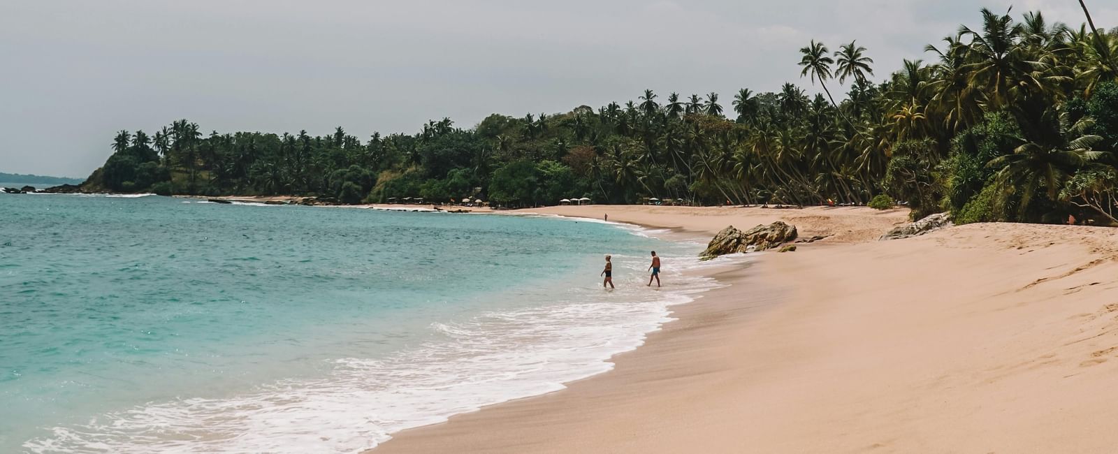 Quiet tropical beach with gentle waves, distant figures walking along shore, and palm trees lining the sandy coastline.