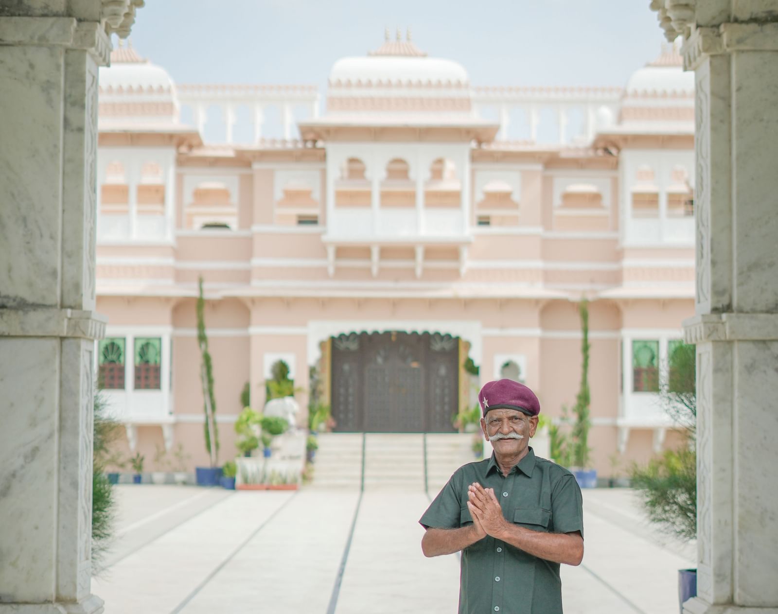A middle-aged man with a mustache and a maroon turban stands facing the camera with his hands pressed together in a traditional greeting at Mahendra Niwas.