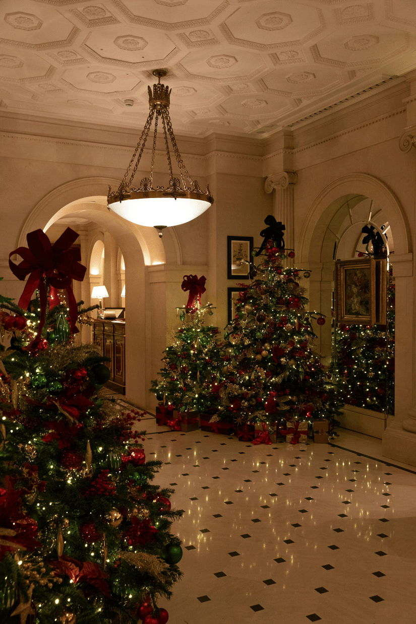 An elegant, dimly lit hall with marble floors featuring three decorated Christmas trees topped with large red bows under a grand chandelier.