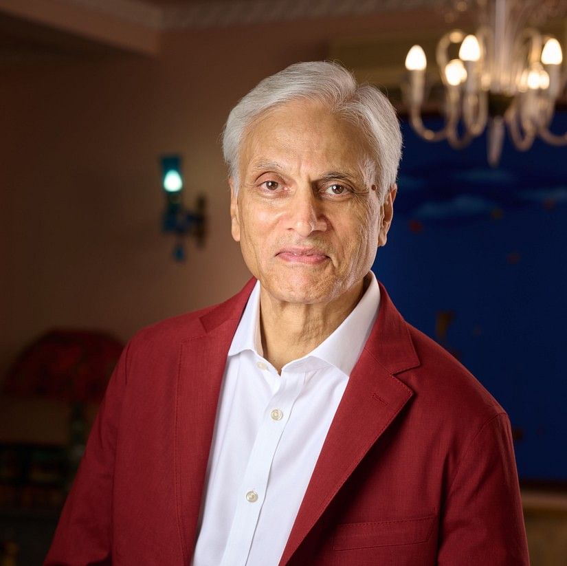 Ajay Bakaya in a red blazer and white shirt, standing with hands clasped, smiling softly in a warm room.