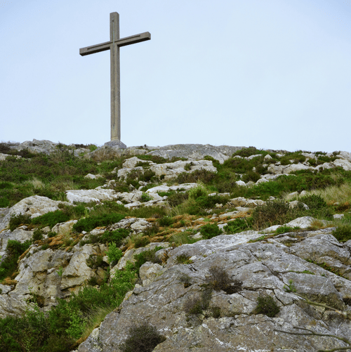A view from below, of the cross above a rocky hill with grass on the hill and blue sky in the background.