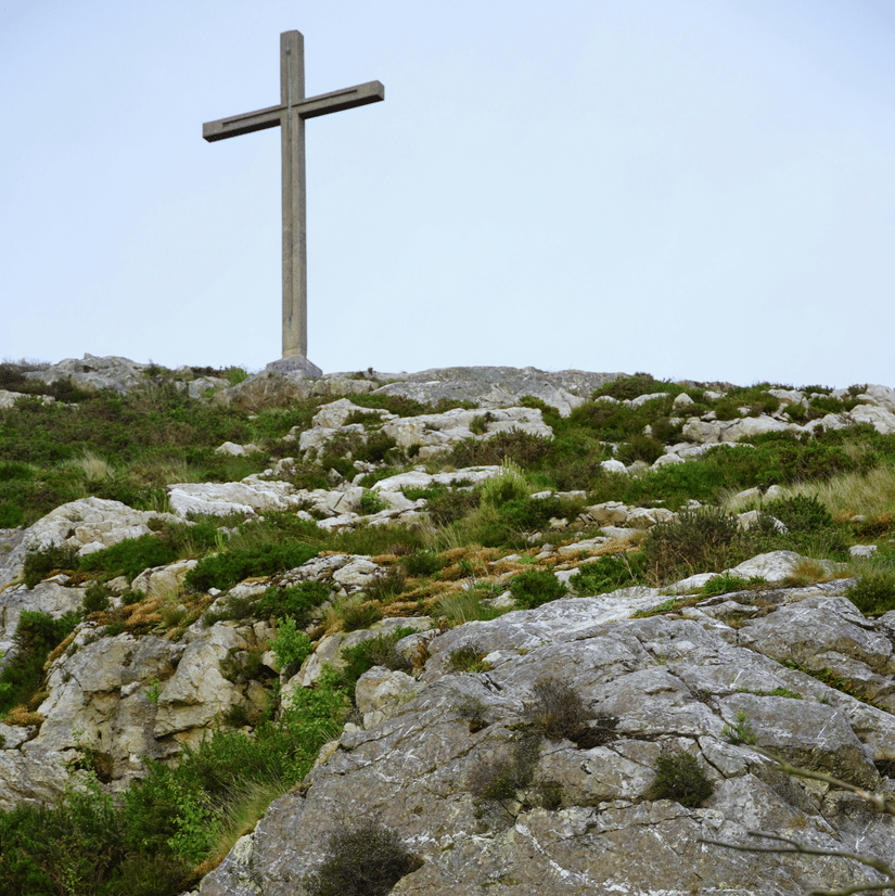 A view from below, of the cross above a rocky hill with grass on the hill and blue sky in the background.