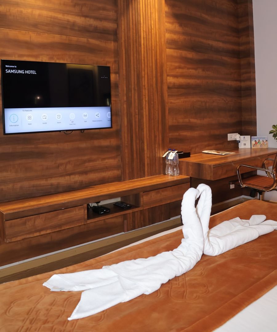 A pair of towel swans on the bed with an LED television mounted on the wooden wall of the Suite at Best Western, Vrindavan.