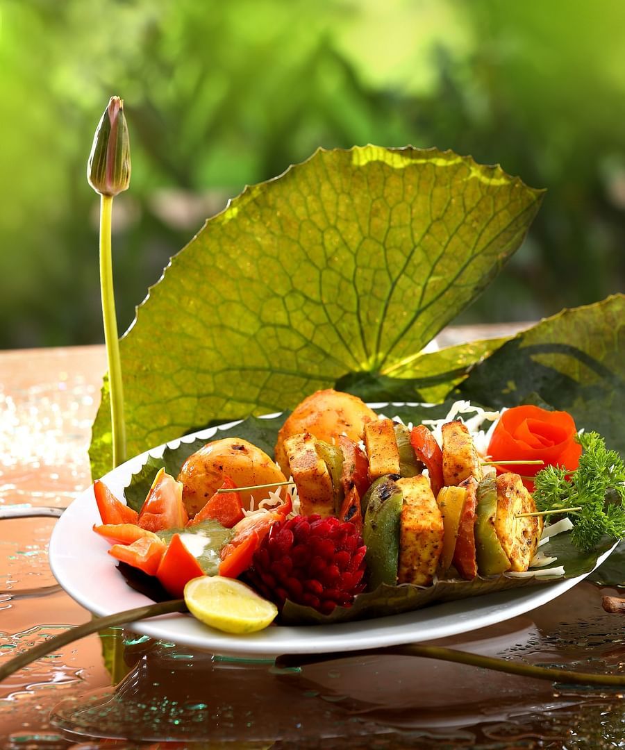 A vegetarian platter sits next to a lotus leaf and bud, while a seafood dish is displayed on the table, with 'Aambal' illuminated on the wall in the background at Uday Backwater Resort