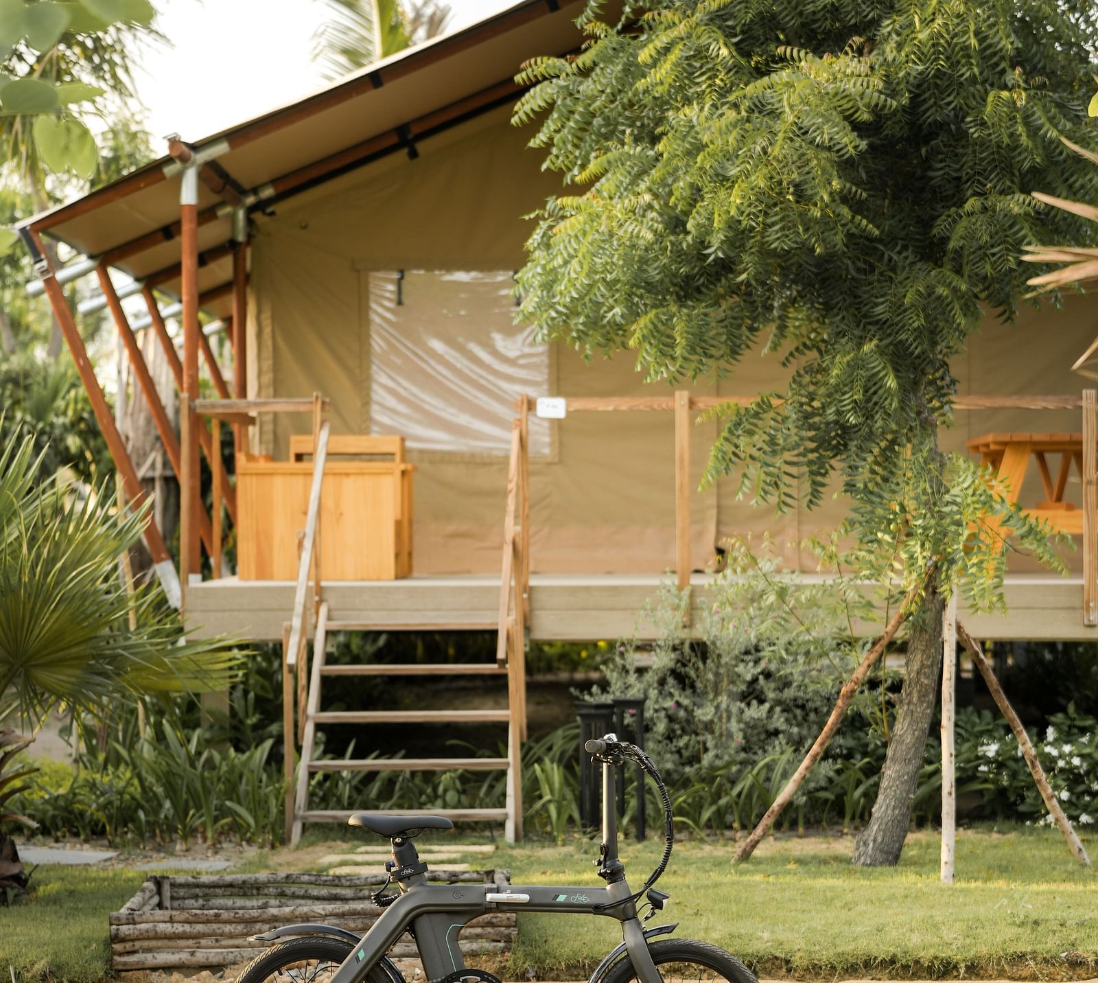 The Peaceful Tent at WoCAL Retreat Ke Ga, seen from across a mud path, featuring an inviting wooden deck and an adventure bicycle parked beside the steps under swaying palm trees.