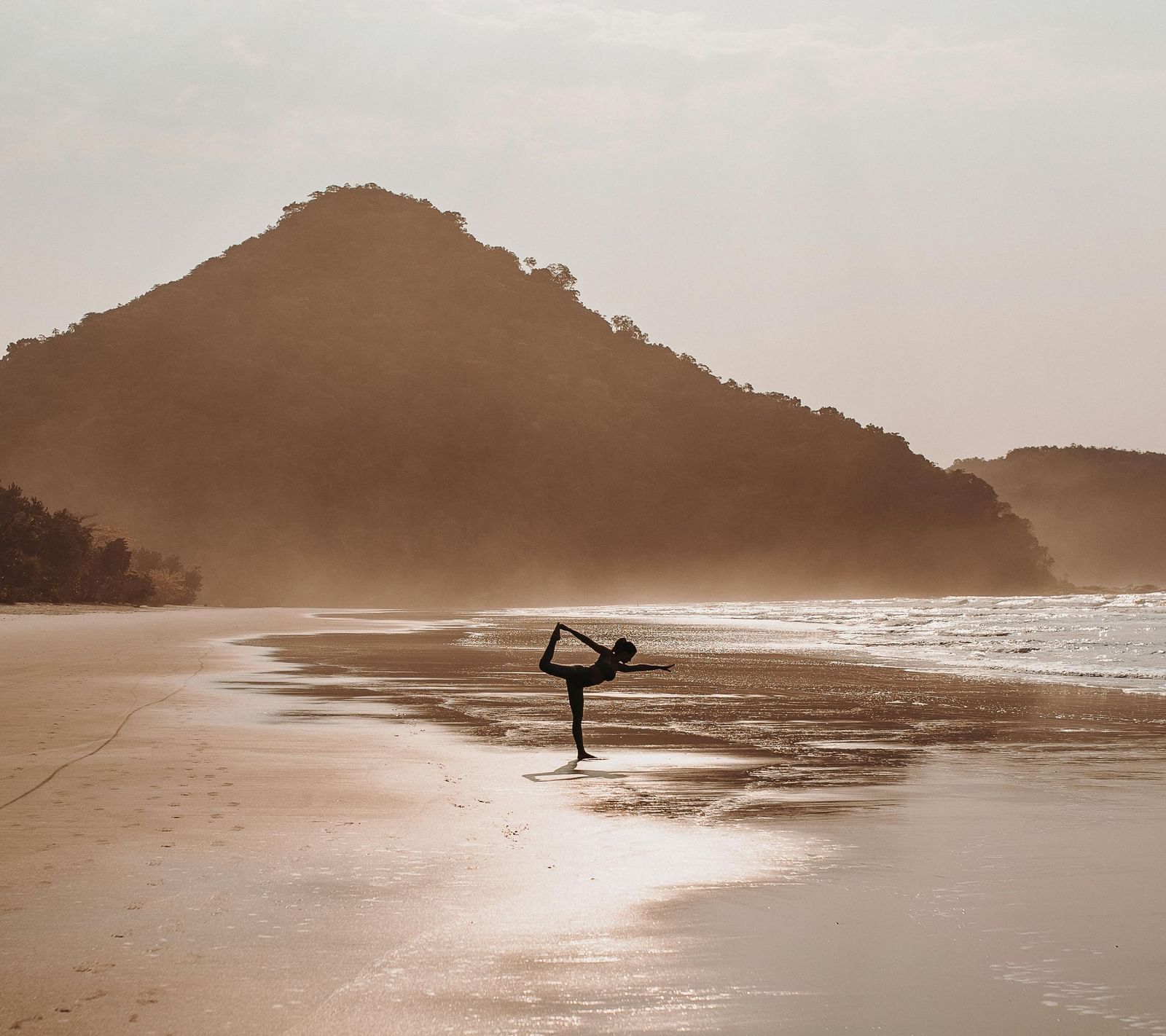 A woman doing yoga on a beach facing towards the sea along with a hill like structure in the background.