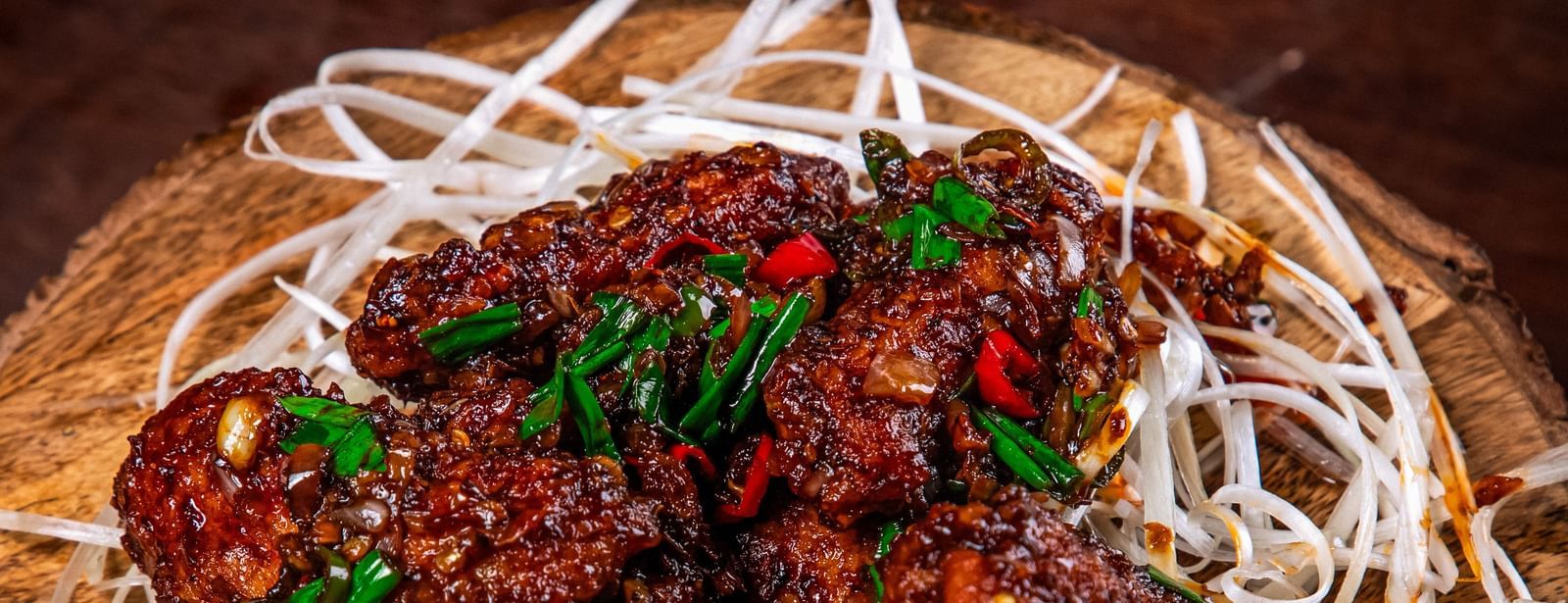 A bowl of crispy, saucy fried chicken strips garnished with sesame seeds and spring onions, placed on shredded white cabbage on a rustic wooden base at The Hamuse Hotel, Kodaikanal.