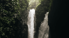 A twin waterfall pours into a dark, rocky cavern from a bright, leaf-framed opening at the top, creating a white pool.