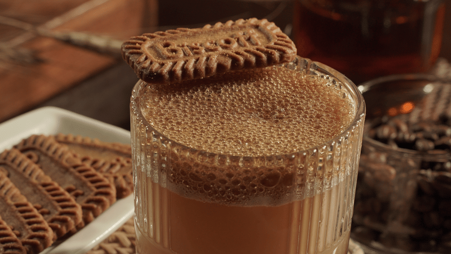 A glass of frothy coffee sits on a woven mat, topped with a cookie. Nearby, more cookies are stacked on a plate, with a background of syrup and coffee beans, creating a cosy, warm atmosphere.