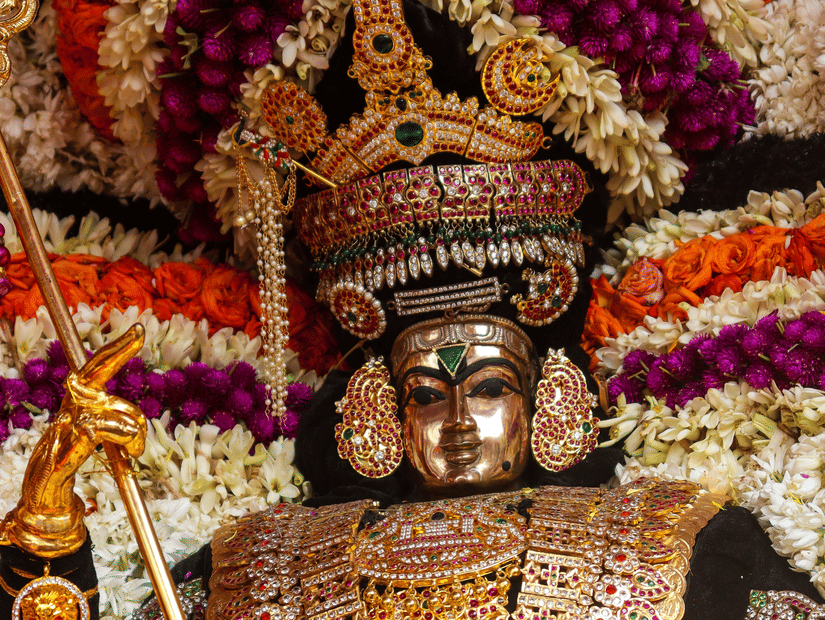 : Close-up of a Hindu deity idol beautifully adorned with heavy gold jewelry, crowns, and vibrant flower garlands.