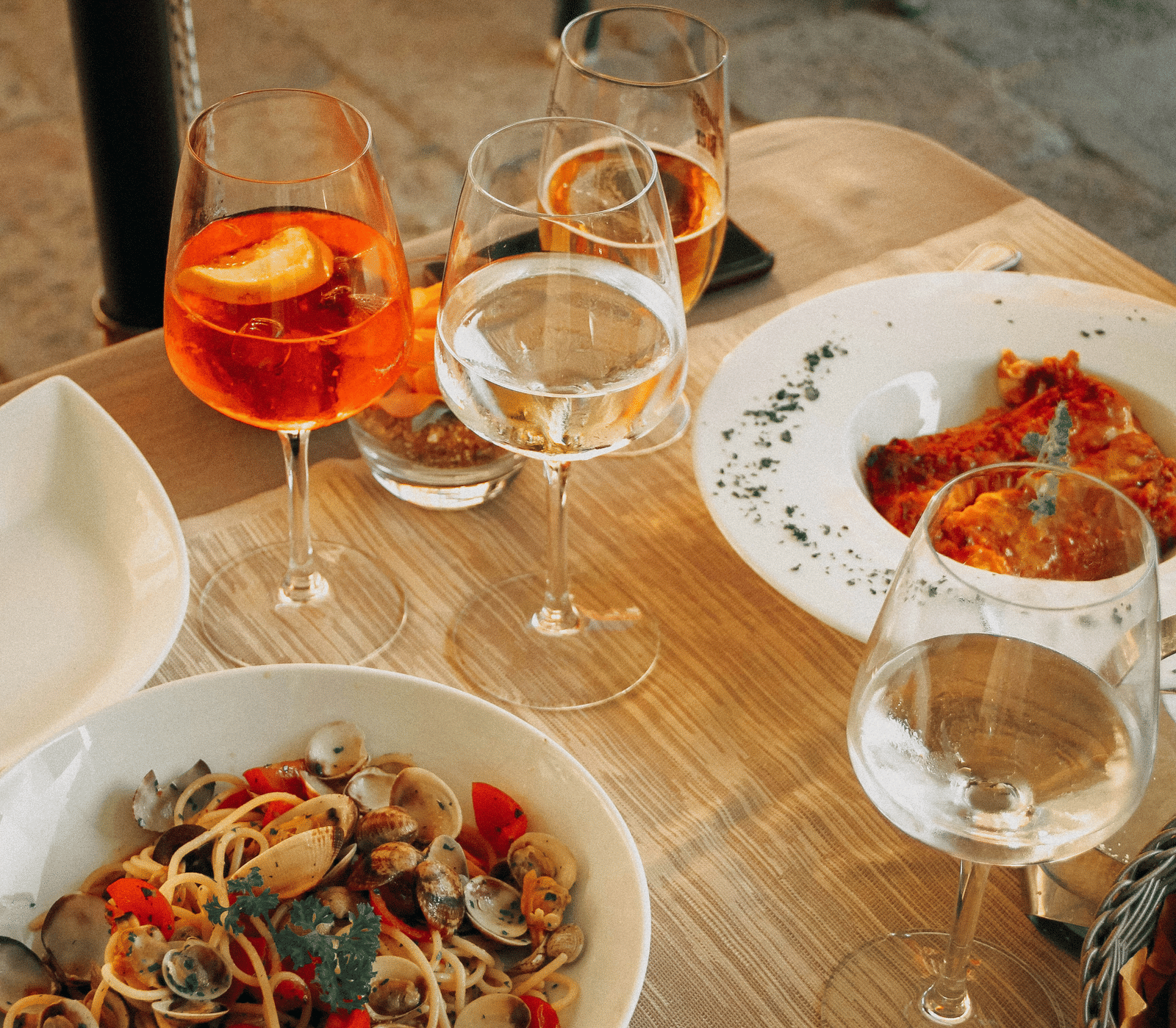 An overhead shot of a wooden table set with bowls of pasta and snacks, along with several glasses of orange and white drinks.