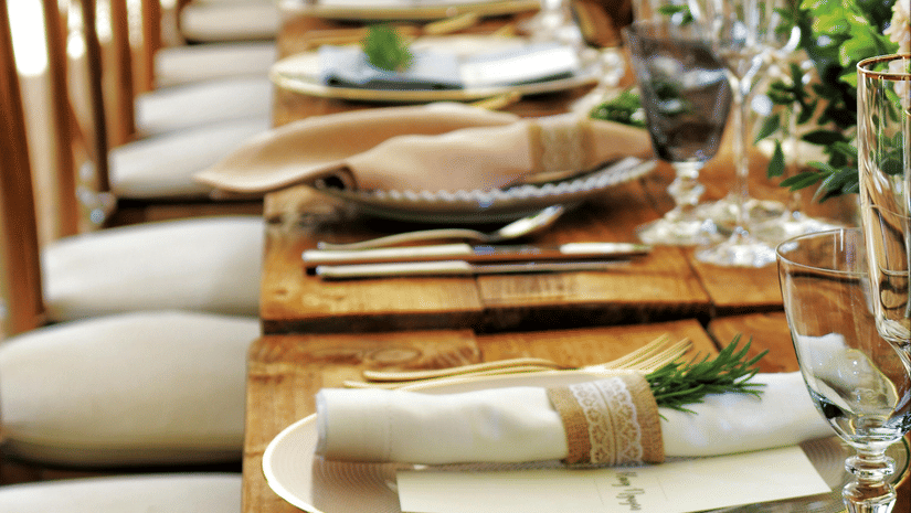 A long wooden table with beautifully set cutlery with small fern leaves in vases.