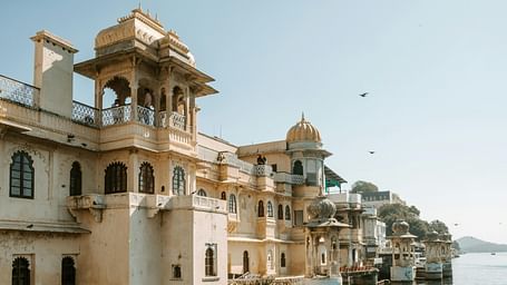 An elegant lakeside view of a historic section of the City Palace in Udaipur, with arched balconies and domed pavilions extending over the tranquil waters of Lake Pichola under a soft morning sky.