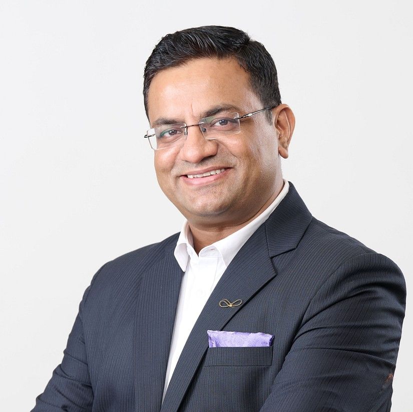 Yash Raghav  in a dark suit with a lavender pocket square, wearing glasses, stands with arms crossed, exuding confidence.