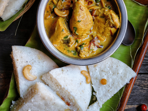 A serving of rice cakes with a bowl of chicken curry served on an earthen plate lined with banana leaf cutout.