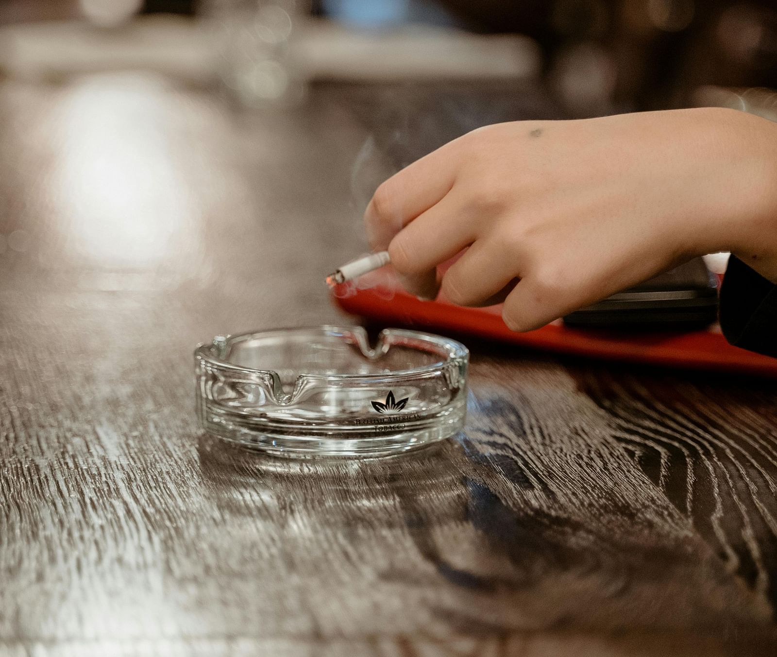 A person holding a cigarette over a crystal ash tray kept on a counter. 