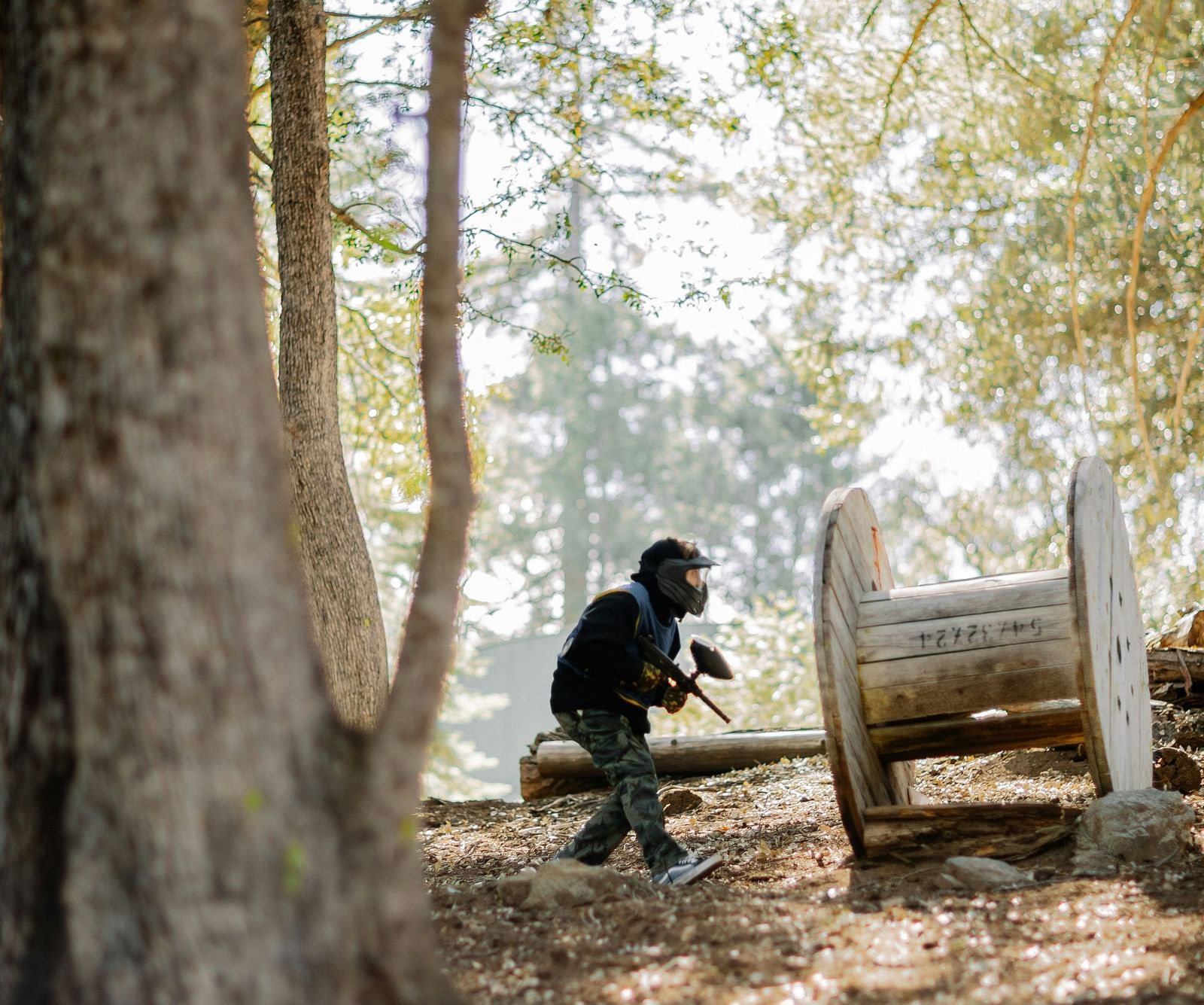 A person in complete paintball gear walking in the field with a giant tree next to him