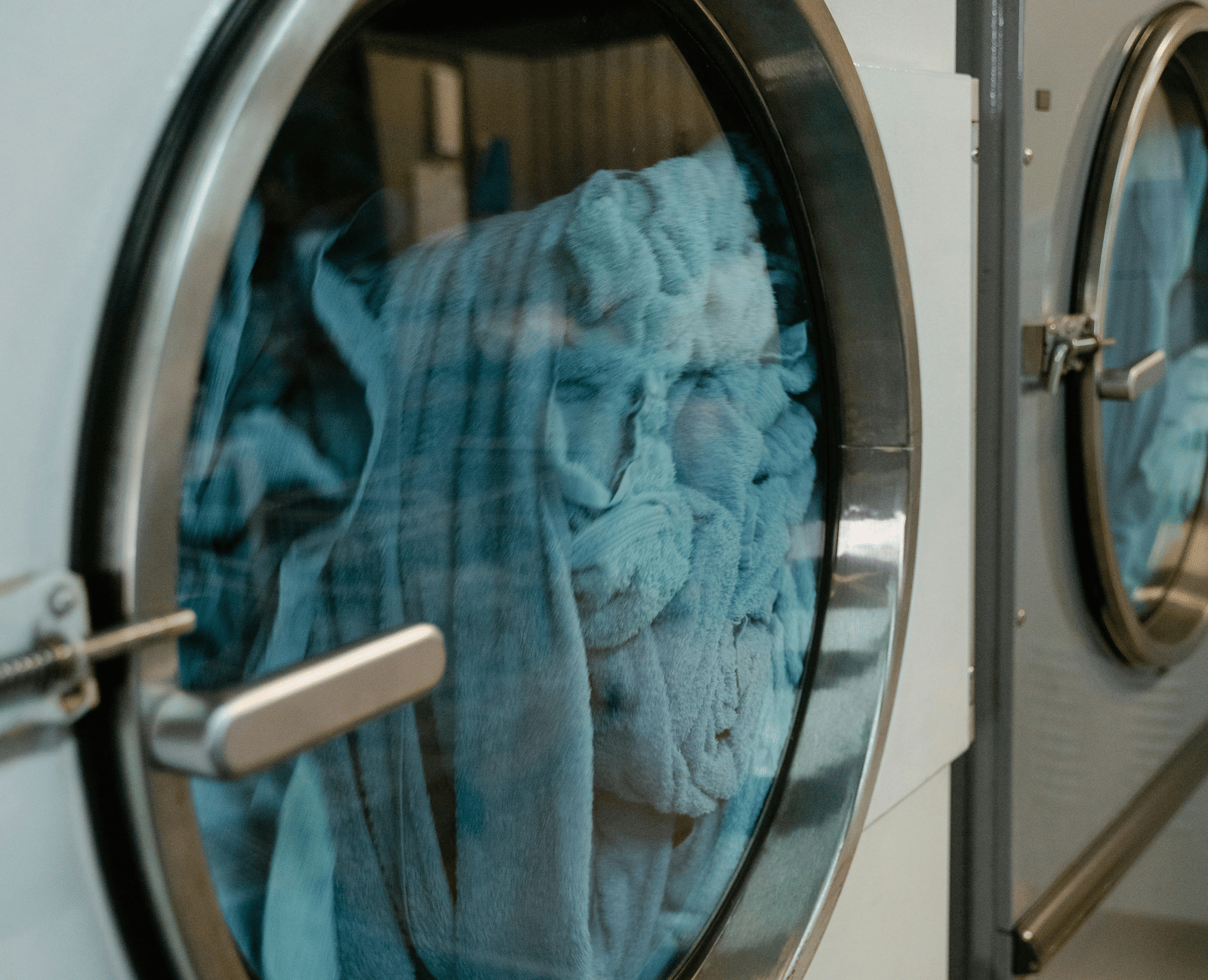 A large industrial tumble dryer door with a round glass window, showing blue laundry inside.