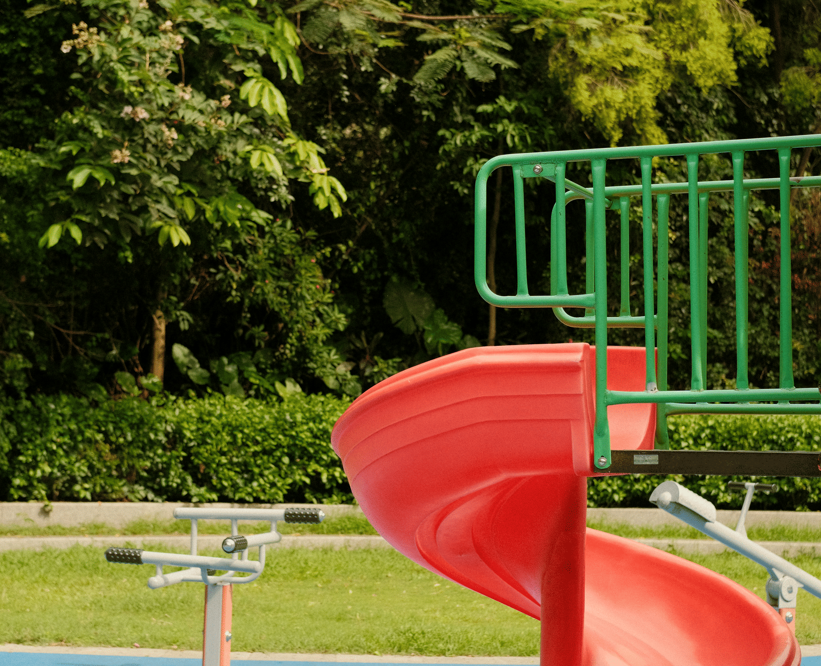 A spiral slide in an outdoor children’s play area with green trees and fencing in the background.