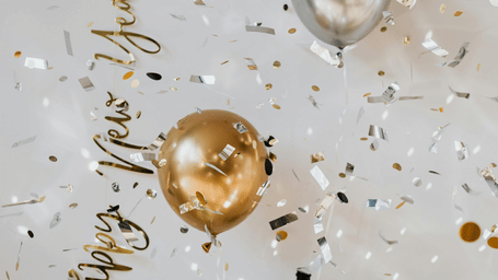 Festive decorations including gold and silver balloons, confetti, and champagne bottles and glasses are arranged on a surface.