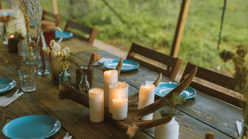 A rustic wooden table set for an outdoor meal under a tent, featuring plates, candles, a dried antler centrepiece, and a forest background.