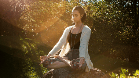 A view where a woman can be seen meditating under sitting in a yoga pose on a rock in a scenic garden filled with green bushes, trees and pink flowers. 