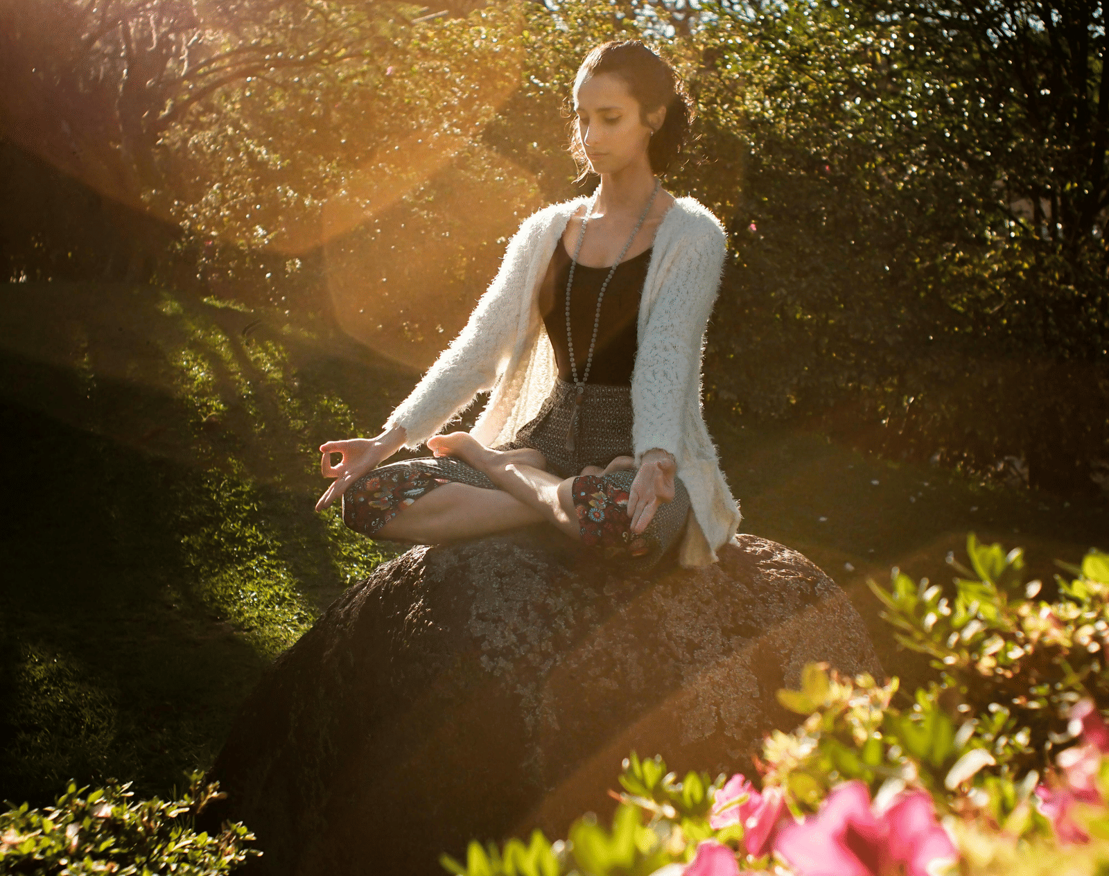 A woman meditating outdoors surrounded by greenery and flowers.